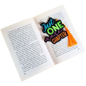 Embroidered 'Just One More Chapter' bookmark on black faux leather placed inside an open book, showing its colourful wording and tassel in use.