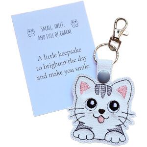 White faux leather kawaii kitty cat keyring with embroidered grey tabby details, shown with a matching keepsake card about charm and smiles.