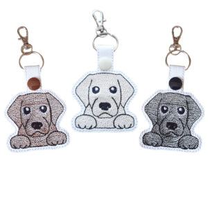Three Kawaii Labrador keyrings in Chocolate, Golden and Black designs, shown with embroidered details and metal hardware.