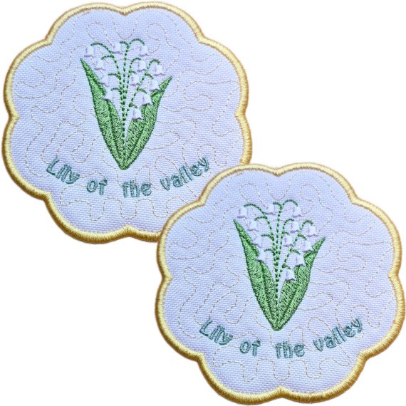 Set of two embroidered May birth flower candle coasters, each with Lily of the Valley flowers, green leaves, stitched text and a scalloped border.