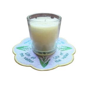 Embroidered May birth flower candle mat with Lily of the Valley flowers, green leaves and a scalloped border, shown underneath a glass candle jar.