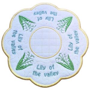 Close up of an embroidered May birth flower candle mat with Lily of the Valley flowers, green leaves, stitched text, background detail and a scalloped border.
