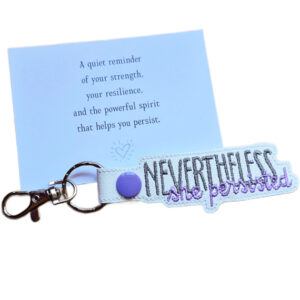 Embroidered Nevertheless She Persisted keyring in white faux leather, shown beside a matching keepsake card with an encouraging message about strength and resilience.