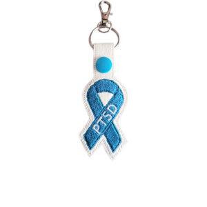 Embroidered PTSD awareness ribbon keyring made from white faux leather, featuring a teal stitched ribbon with PTSD lettering and silver metal keyring hardware.