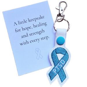 Embroidered PTSD awareness ribbon keyring made from white faux leather with a teal stitched ribbon, shown beside a keepsake card with a message of hope, healing and strength.