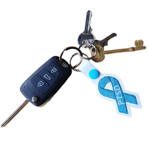 Embroidered PTSD awareness ribbon keyring attached to a set of keys, showing the teal stitched ribbon design on white faux leather with silver metal hardware.