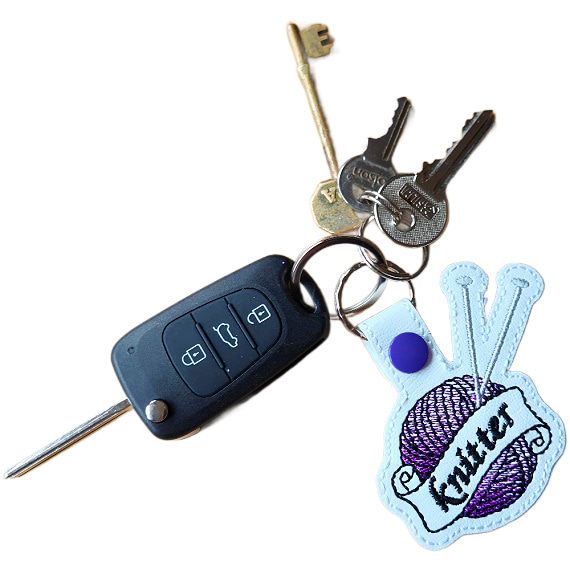 White faux leather knitting keyring attached to a set of keys, embroidered with a purple yarn ball, silver knitting needles and banner reading 'Knitter'.