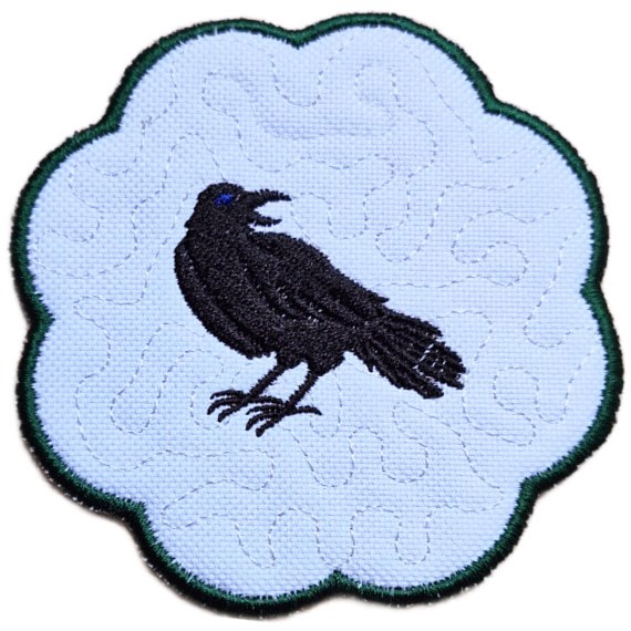 Close up of an embroidered raven candle coaster with a striking black raven, scalloped edging and subtle gothic background detail.