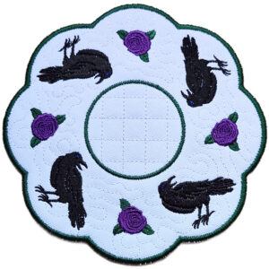 Close up of an embroidered raven candle mat showing black ravens, purple rose details, scalloped edging and a central circle for a candle or display item.