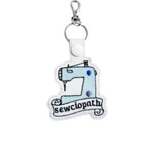 White faux leather sewing machine keyring embroidered with blue detail and the word 'Sewciopath', finished with a metal clasp.