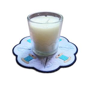 Embroidered sewing candle mat with stitched thread spools, flowing thread details, scalloped edging and a glass jar candle displayed in the centre.