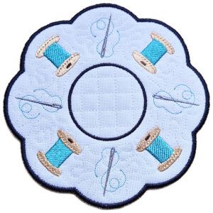 Close up of an embroidered sewing candle mat showing stitched thread spools, flowing needle and thread details, scalloped edging and a central circle for a candle or display item.