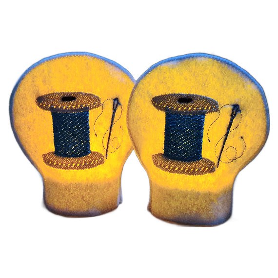 Lit sewing tealight covers glowing warmly over LED tealights, showing embroidered thread spool designs with needle and thread details for a cosy craft room display.