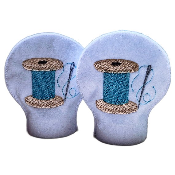 Pair of unlit embroidered sewing tealight covers made from soft felt, showing stitched thread spool designs with needle and thread details for a cosy craft room display.