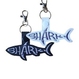 Black and white faux leather shark word art keyrings with grey embroidered 'Shark' lettering, snap fastenings and silver lobster clasp hardware.