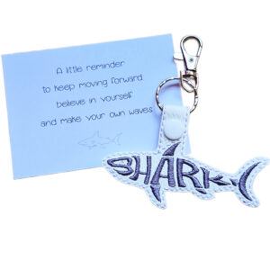 White faux leather shark word art keyring with grey embroidered lettering, shown beside a matching white keepsake card with ocean-themed wording.