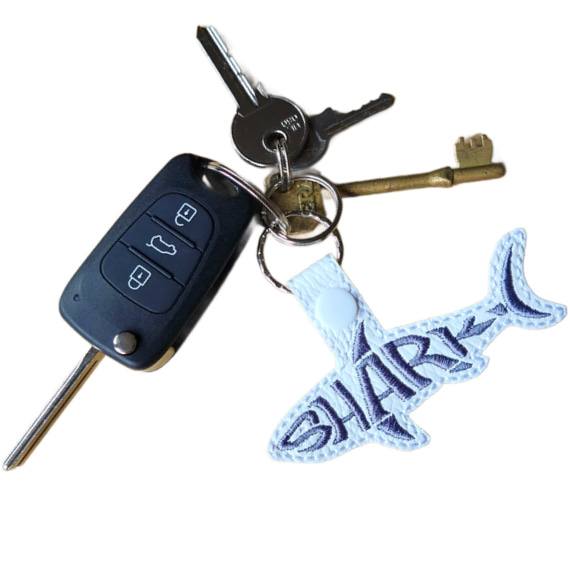 White faux leather shark word art keyring with grey embroidered 'Shark' lettering, shown attached to a set of keys with silver keyring hardware.