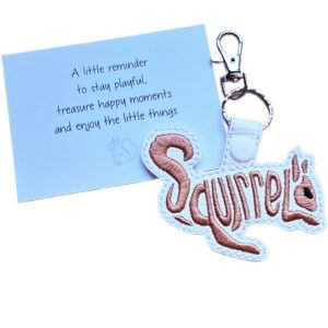 White faux leather squirrel word art keyring with brown embroidered 'Squirrel' lettering, shown beside a white keepsake card with woodland-themed wording.
