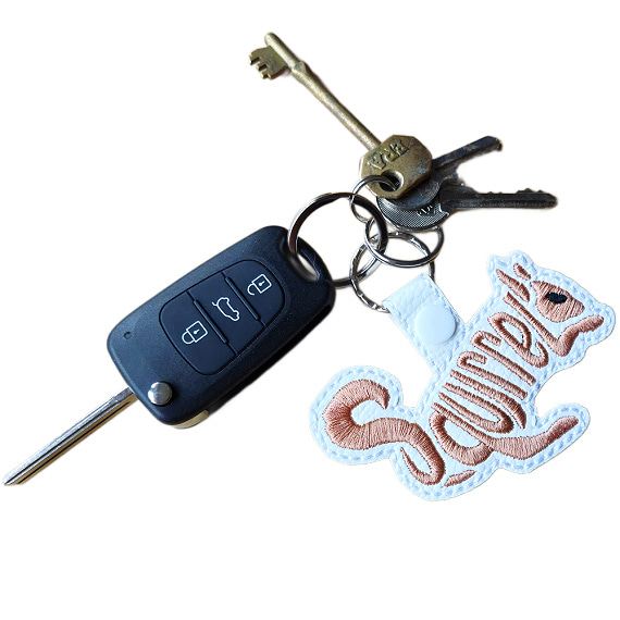 White faux leather squirrel word art keyring with brown embroidered 'Squirrel' lettering, shown attached to a set of keys with silver keyring hardware.