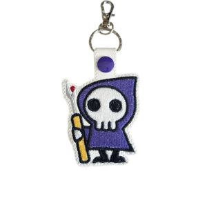 White faux leather Stitch Reaper keyring with a purple cloak and seam ripper, shown with a keepsake card.