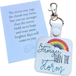 White faux leather Stronger Than the Storm keyring with embroidered rainbow and cloud design, shown beside a white keepsake card with hopeful reminder wording.