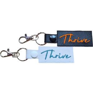 Embroidered Thrive keyring in faux leather, with the word 'Thrive' stitched on black and white backgrounds with sturdy metal hardware.