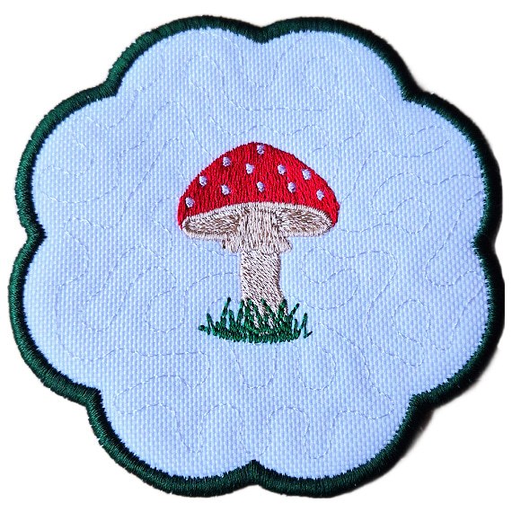 Close up of an embroidered toadstool candle coaster with a red stitched toadstool, grassy detail, scalloped edging and subtle woodland background stitching.