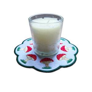 Embroidered toadstool candle mat with red stitched toadstools, grassy details, scalloped edging and a glass jar candle displayed in the centre.