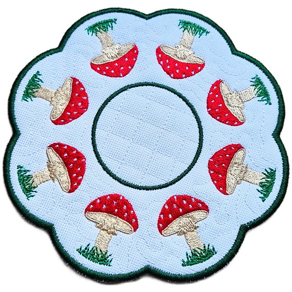 Close up of an embroidered toadstool candle mat showing red stitched toadstools, grassy details, scalloped edging and a central circle for a candle or display item.