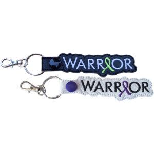Embroidered Warrior awareness ribbon keyrings shown in black and white faux leather, each with bold stitched wording, coloured ribbon detail and metal keyring hardware.