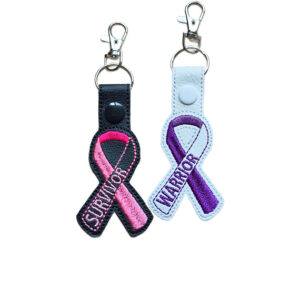 Embroidered Awareness Ribbon keyrings shown in black and white faux leather, featuring Survivor and Warrior designs with pink and purple stitched ribbon details.
