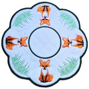 Close up of an embroidered woodland fox candle mat with orange foxes, green grass details, circular centre and dark green scalloped border.
