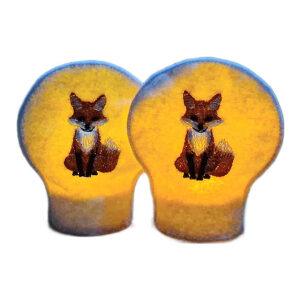 Two lit woodland fox tealight covers glowing warmly over LED tealights, each embroidered with a cosy orange fox on white felt.
