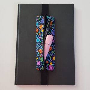 Handmade A4 planner zipper pouch in colourful spot print cotton fabric, shown fitted around a black A4 notebook with a black zip and elastic strap, holding pens and small stationery items.