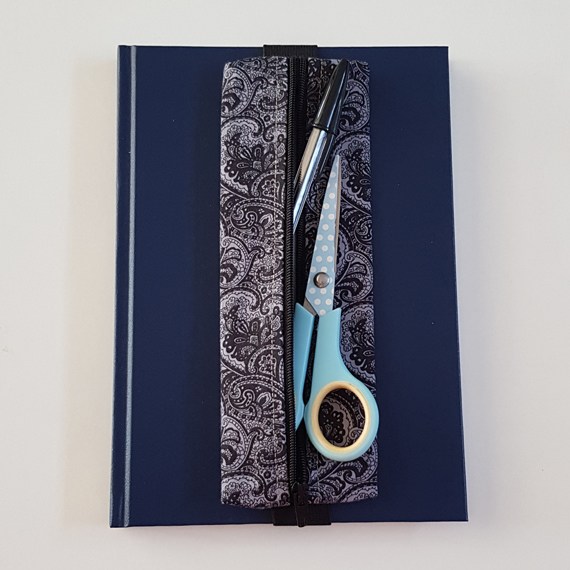 Handmade A5 planner zipper pouch in black and white paisley print cotton fabric, shown fitted around a navy A5 notebook with a black zip and elastic strap, holding pens and small stationery items.