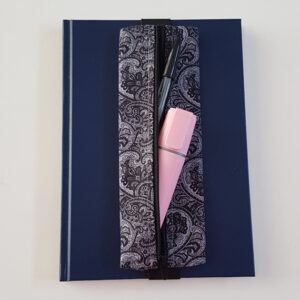 Handmade A5 planner zipper pouch in black and white paisley print cotton fabric, shown fitted around a navy A5 notebook with a black zip and elastic strap, holding pens and small stationery items.