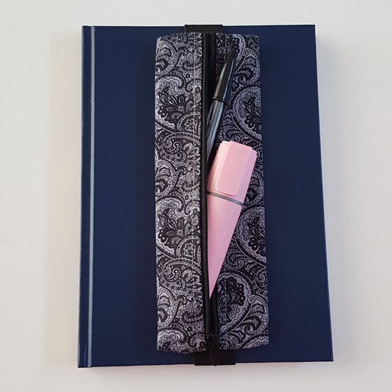 Handmade A5 planner zipper pouch in black and white paisley print cotton fabric, shown fitted around a navy A5 notebook with a black zip and elastic strap, holding pens and small stationery items.