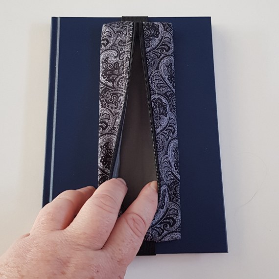 Handmade A5 planner zipper pouch in black and white paisley print cotton fabric, shown fitted around a navy A5 notebook with a black zip and elastic strap, holding pens and small stationery items.