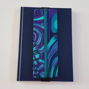 Handmade A5 planner zipper pouch made from teal, turquoise, blue and purple paua shell print cotton fabric, shown fitted around a navy A5 notebook with a zipped compartment for pens and stationery.
