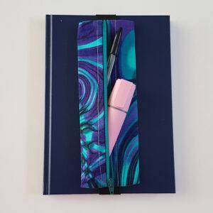 Handmade A5 planner zipper pouch made from teal, turquoise, blue and purple paua shell print cotton fabric, shown fitted around a navy A5 notebook with a zipped compartment for pens and stationery.
