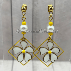 Handmade floral drop earrings featuring white enamel flower charms within gold geometric frames, accented with pearl beads and yellow details, creating elegant lightweight jewellery for spring and summer wear.