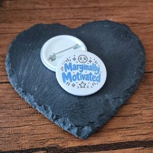 Marginally Motivated funny mood badge with playful blue lettering, star accents and a small unimpressed face, available as a badge, magnet or keyring.
