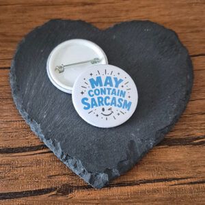 Funny sarcastic badge design with the phrase 'May contain sarcasm' in bold playful lettering, decorated with small sparkle and starburst accents.