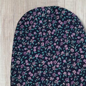 Handmade purple floral stoma bag cover with small purple flowers, teal green leaves and dark navy fabric, shown close up on a wooden background.