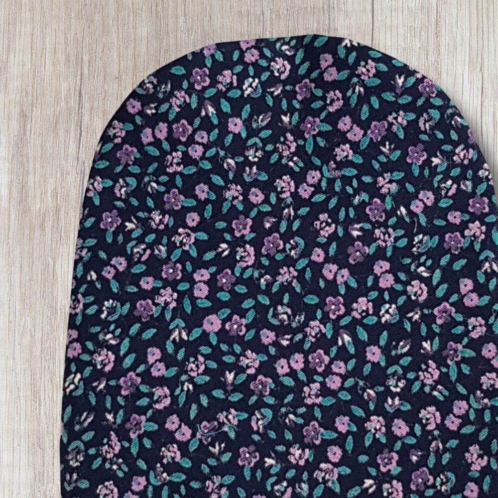 Handmade purple floral stoma bag cover with small purple flowers, teal green leaves and dark navy fabric, shown close up on a wooden background.